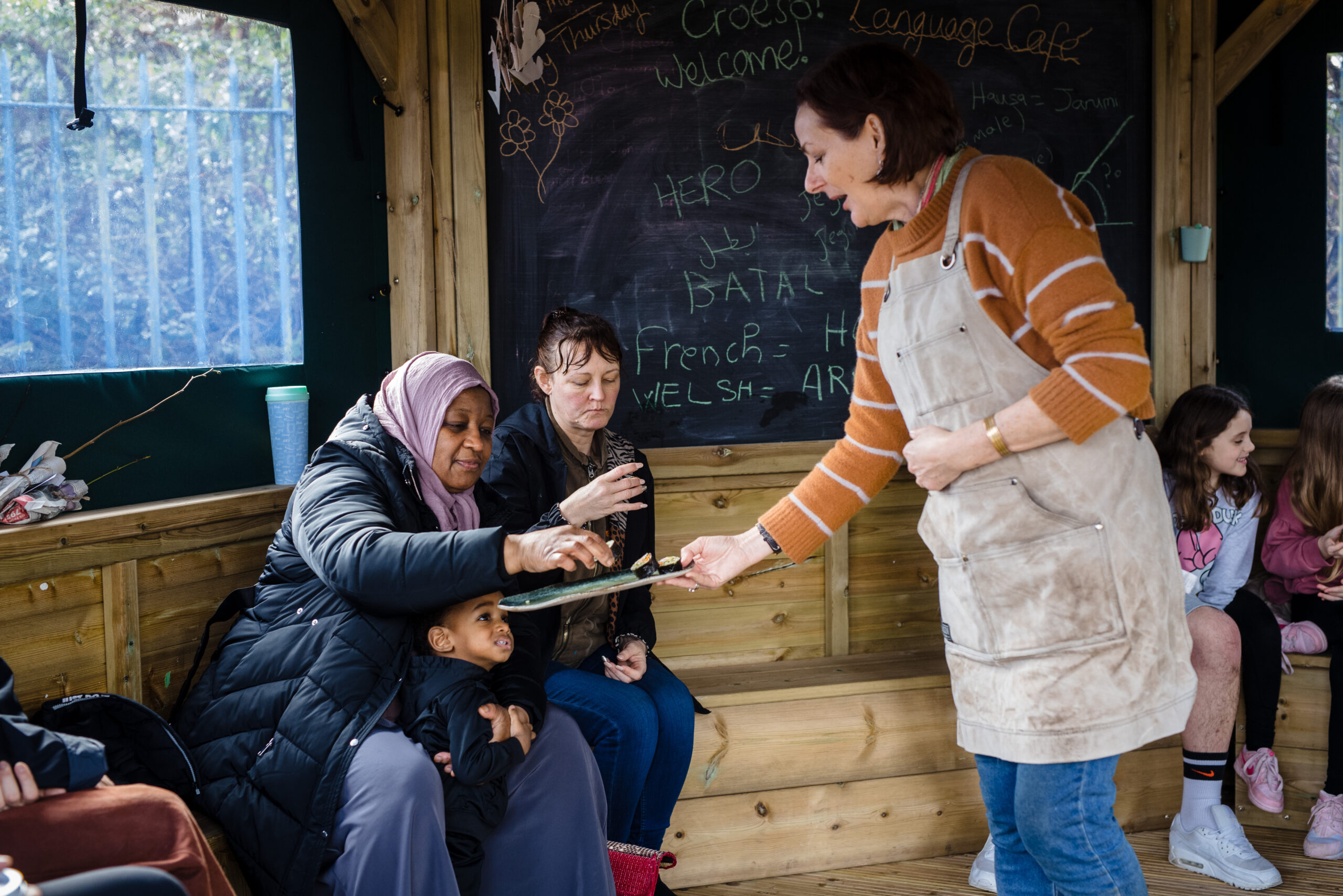 A picture of a white woman with an orange jumper and a beige apron offering food to two other women, who are both sitting down, and a young boy. The woman on the left is wearing a lilac hijab and a black coat and is smiling peacefully. The woman on the right is a white woman with brown hair, wearing a black coat and blue jeans. The young boy is standing between the legs of the woman with a hijab and is looking up at the tray. All are standing in a wooden-panelled room, decorated with a black board.